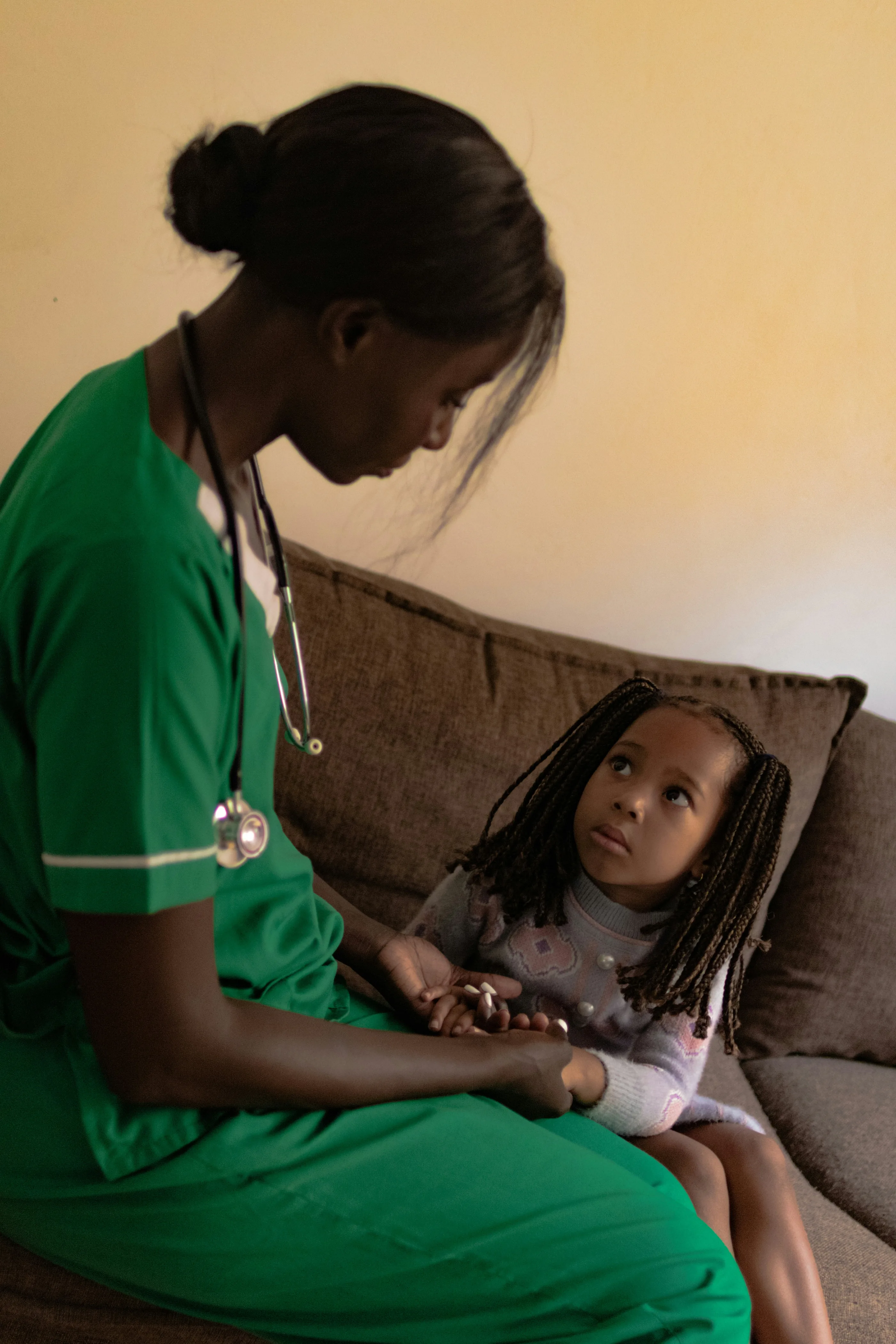 A nurse in green scrubs sits with a young girl, who looks up at her.